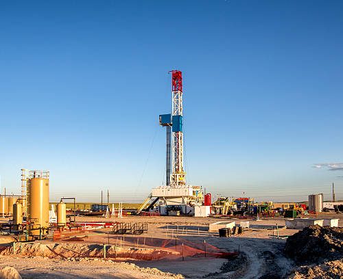 A lone drilling Fracking Rig set against the dramatic landscape of the American Prairie lands.  The sun is setting just off camera creating beautiful yellows and oranges on the clouds above.  In the far distance, wind turbines are visible.